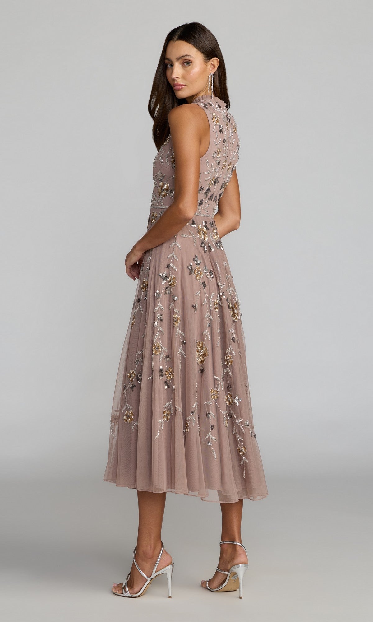 Wearing the Mac Duggal 10815 midi wedding guest dress—sleeveless, blush pink with a high neckline, floral sequin embroidery, and an elegant a-line skirt—a woman pairs it with silver high heels as she stands on a plain backdrop, looking over her shoulder.